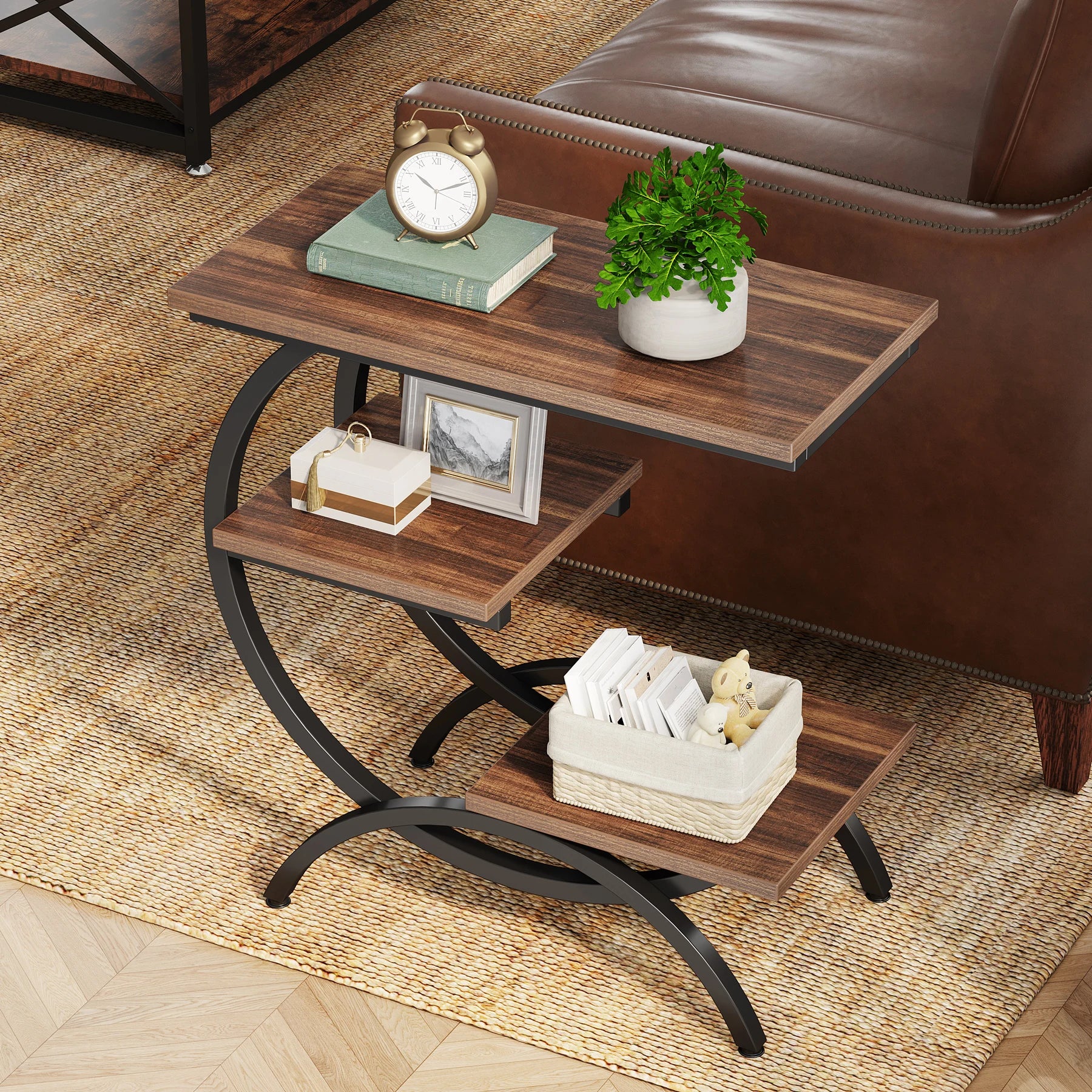 Compact 3-tier C-shape end table with wood shelves and black metal frame, styled with books and a clock beside a leather sofa.